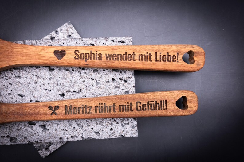 Zwei gravierte Kochlöffel aus Akazienholz mit den Gravuren „Sophia wendet mit Liebe!“ und „Moritz rührt mit Gefühl!!!“. Beide Griffe sind mit Herz-Ausschnitt und Symbolen versehen. Die präzise Lasergravur hebt sich kontrastreich vom warmen Holzton ab. Perfektes personalisierbares Geschenk für Hobbyköche, Paare oder Freunde, die gerne gemeinsam kochen.