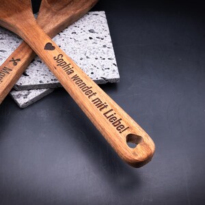 Nahaufnahme des personalisierten Akazienholz-Kochlöffelsets mit Gravur „Sophia wendet mit Liebe!“ und Herzsymbol. Die dunkle Gravur hebt sich kontrastreich vom warmen Holzton ab. Ideal zur individuellen Gestaltung mit Namen, Symbol oder Spruch – ein originelles Geschenk für Küche, Familie oder Freunde.
