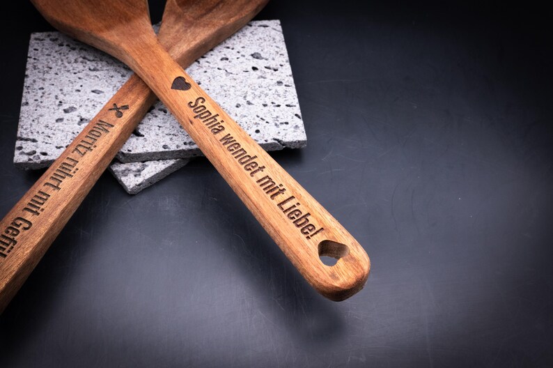 Nahaufnahme des personalisierten Akazienholz-Kochlöffelsets mit Gravur „Sophia wendet mit Liebe!“ und Herzsymbol. Die dunkle Gravur hebt sich kontrastreich vom warmen Holzton ab. Ideal zur individuellen Gestaltung mit Namen, Symbol oder Spruch – ein originelles Geschenk für Küche, Familie oder Freunde.