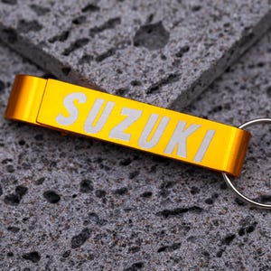 Goldfarbener Flaschenöffner Schlüsselanhänger aus Aluminium mit Gravur „SUZUKI“ in weißer Schrift. Praktischer Schlüsselanhänger mit integriertem Kapselheber, robust und leicht, ideal für personalisierte Gravuren mit Namen, Logos oder Text. Elegantes Geschenk oder Werbeartikel für Autofans, Motorradfahrer und Markenliebhaber, fotografiert auf grauem Steinuntergrund.
