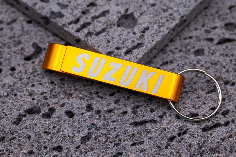 Goldfarbener Flaschenöffner Schlüsselanhänger aus Aluminium mit Gravur „SUZUKI“ in weißer Schrift. Praktischer Schlüsselanhänger mit integriertem Kapselheber, robust und leicht, ideal für personalisierte Gravuren mit Namen, Logos oder Text. Elegantes Geschenk oder Werbeartikel für Autofans, Motorradfahrer und Markenliebhaber, fotografiert auf grauem Steinuntergrund.