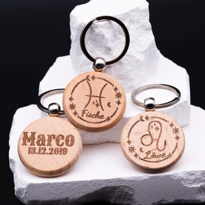 Drei gravierte Holz-Schlüsselanhänger mit personalisierbaren Sternzeichen liegen auf gestapeltem weißen Stein. Zu sehen sind Fische mit Sternkreisgravur, Löwe sowie ein Anhänger mit individueller Gravur „Marco 13.12.2019“. Jeder Anhänger hat einen stabilen Schlüsselring aus Metall. Die Kombination aus natürlichem Holz, feiner Lasergravur und astrologischen Symbolen macht diese Schlüsselanhänger zu einem persönlichen Geschenk mit individuellem Charakter.