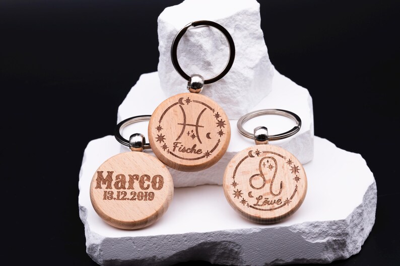 Drei gravierte Holz-Schlüsselanhänger mit personalisierbaren Sternzeichen liegen auf gestapeltem weißen Stein. Zu sehen sind Fische mit Sternkreisgravur, Löwe sowie ein Anhänger mit individueller Gravur „Marco 13.12.2019“. Jeder Anhänger hat einen stabilen Schlüsselring aus Metall. Die Kombination aus natürlichem Holz, feiner Lasergravur und astrologischen Symbolen macht diese Schlüsselanhänger zu einem persönlichen Geschenk mit individuellem Charakter.