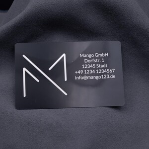 Visitenkarte aus schwarzem Aluminium mit Gravur des Mango-Digitals-Logos und vollständigen Kontaktdaten (Adresse, Telefon, E-Mail). Das Design ist modern und elegant, die Metallkarte robust und langlebig. Ideal als exklusive Alternative zur klassischen Papierkarte, perfekt für professionelle Business-Präsentationen und Networking.