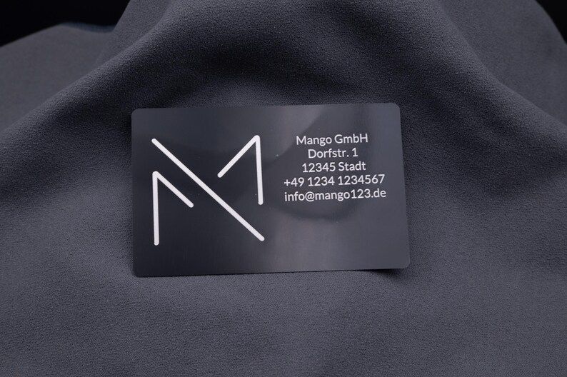 Visitenkarte aus schwarzem Aluminium mit Gravur des Mango-Digitals-Logos und vollständigen Kontaktdaten (Adresse, Telefon, E-Mail). Das Design ist modern und elegant, die Metallkarte robust und langlebig. Ideal als exklusive Alternative zur klassischen Papierkarte, perfekt für professionelle Business-Präsentationen und Networking.