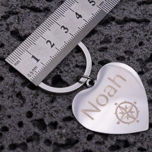 Edelstahl-Herzanhänger mit Gravur des Namens „Noah“ und Kompasssymbol, neben einem Metalllineal zur Längenmessung. Der Schlüsselanhänger kombiniert glänzendes Material mit klarer Gravur und symbolischer Bedeutung. Perfekt als stilvolles Accessoire oder Geschenkidee für Reisende, Partner oder Freunde.