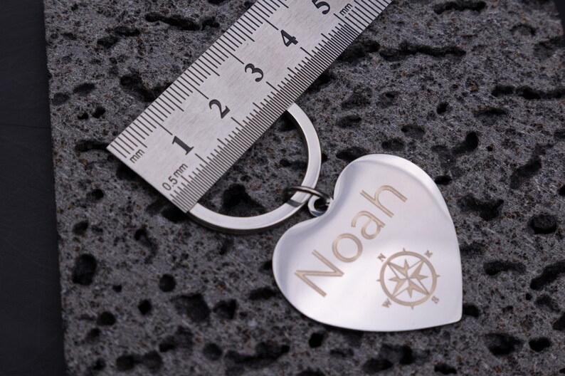 Edelstahl-Herzanhänger mit Gravur des Namens „Noah“ und Kompasssymbol, neben einem Metalllineal zur Längenmessung. Der Schlüsselanhänger kombiniert glänzendes Material mit klarer Gravur und symbolischer Bedeutung. Perfekt als stilvolles Accessoire oder Geschenkidee für Reisende, Partner oder Freunde.