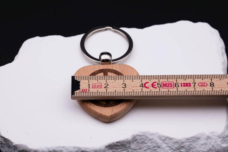 Herz Schlüsselanhänger aus Holz mit Peace-Symbol und Maßband zur Darstellung der Größe. Personalisierbar mit individuellen Gravuren wie Namen oder Symbolen, ist dieser Anhänger ein besonderes Geschenk für Familie, Freunde oder Partner. Hergestellt aus hochwertigem Naturholz, modern graviert und langlebig. Perfekt als Geschenkidee für Geburtstag, Jubiläum, Hochzeitstag oder für den Alltag.