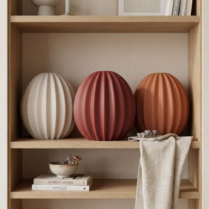 May include: Three decorative ceramic spheres in cream, red-orange, and terracotta hues, each with vertical ribbed detailing. The spheres are displayed on a light wood shelf alongside books, a small bowl, and a folded linen cloth.
