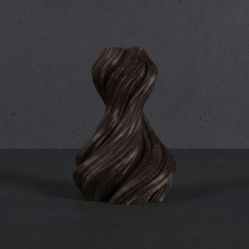 K&ouml;nnte beinhalten: Eine dunkelbraune, dekorative Vase mit einem einzigartigen, gedrehten Design. Die Vase hat eine strukturierte Oberfl&auml;che mit feinen Sprenkeln, und die Form erinnert an flie&szlig;ende B&auml;nder. Ein stilvolles Wohnaccessoire.