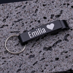 Schwarzer Flaschenöffner Schlüsselanhänger aus Aluminium mit Gravur „Emilia“ und Herz-Symbol. Der praktische Kapselheber mit Schlüsselring ist stabil, leicht und personalisierbar. Perfekt als Geschenk oder individuelles Accessoire für den Schlüsselbund, verbindet Funktionalität mit persönlicher Note. Präsentiert auf grauem Steinuntergrund für eine moderne Produktdarstellung.
