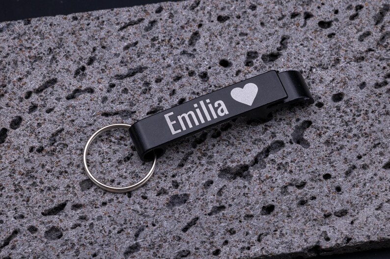 Schwarzer Flaschenöffner Schlüsselanhänger aus Aluminium mit Gravur „Emilia“ und Herz-Symbol. Der praktische Kapselheber mit Schlüsselring ist stabil, leicht und personalisierbar. Perfekt als Geschenk oder individuelles Accessoire für den Schlüsselbund, verbindet Funktionalität mit persönlicher Note. Präsentiert auf grauem Steinuntergrund für eine moderne Produktdarstellung.