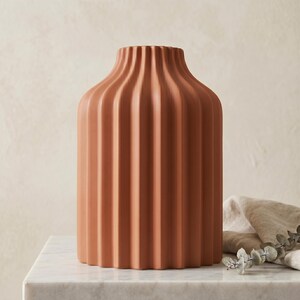 Może przedstawiać: Ceramiczny wazon w kolorze terakoty z żebrowanym wzorem. Wazon ma cylindryczny korpus i lekko rozszerzającą się szyję. Wazon stoi na białej marmurowej powierzchni.