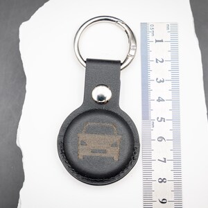 Schwarzer AirTag Schlüsselanhänger aus PU Leder mit integrierter Halterung, präsentiert auf weißem Stein mit danebenliegendem Lineal zur Größenveranschaulichung. Die Vorderseite zeigt eine dezente Lasergravur in Form eines Autosymbols. Der stabile Schlüsselring aus Metall ist oben befestigt. Das Bild verdeutlicht die kompakten Abmessungen des Anhängers sowie die passgenaue Form für den Apple AirTag. Ideal als personalisierter Schlüsselanhänger für Auto und Schlüsselbund.
