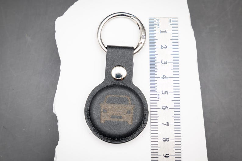 Schwarzer AirTag Schlüsselanhänger aus PU Leder mit integrierter Halterung, präsentiert auf weißem Stein mit danebenliegendem Lineal zur Größenveranschaulichung. Die Vorderseite zeigt eine dezente Lasergravur in Form eines Autosymbols. Der stabile Schlüsselring aus Metall ist oben befestigt. Das Bild verdeutlicht die kompakten Abmessungen des Anhängers sowie die passgenaue Form für den Apple AirTag. Ideal als personalisierter Schlüsselanhänger für Auto und Schlüsselbund.