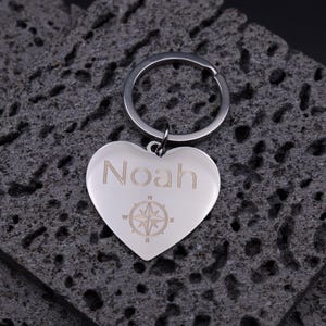 Edler Herz-Schlüsselanhänger aus glänzendem Edelstahl mit Gravur des Namens „Noah“ und einer Kompassrose. Der Anhänger liegt auf dunklem Steinuntergrund und reflektiert sanft das Licht. Die feine Lasergravur hebt sich dezent vom polierten Metall ab und verleiht dem Schlüsselanhänger eine persönliche, hochwertige Note – ideal als Geschenk für Partner oder Freunde.