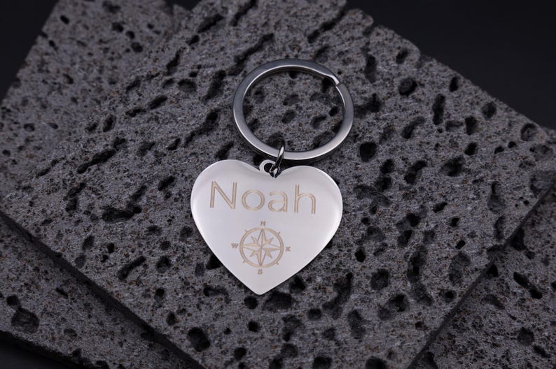 Edler Herz-Schlüsselanhänger aus glänzendem Edelstahl mit Gravur des Namens „Noah“ und einer Kompassrose. Der Anhänger liegt auf dunklem Steinuntergrund und reflektiert sanft das Licht. Die feine Lasergravur hebt sich dezent vom polierten Metall ab und verleiht dem Schlüsselanhänger eine persönliche, hochwertige Note – ideal als Geschenk für Partner oder Freunde.