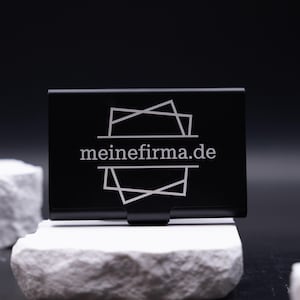 Schwarzes Visitenkartenetui aus Metall mit Lasergravur „meinefirma.de“ auf der Vorderseite. Modernes, minimalistisches Design auf weißem Stein fotografiert. Das Etui ist leicht, robust und perfekt für Geschäftsleute, die ihre Visitenkarten stilvoll und professionell präsentieren möchten. Die Gravur macht es zu einem personalisierten Accessoire für Büro, Meetings oder Networking.
