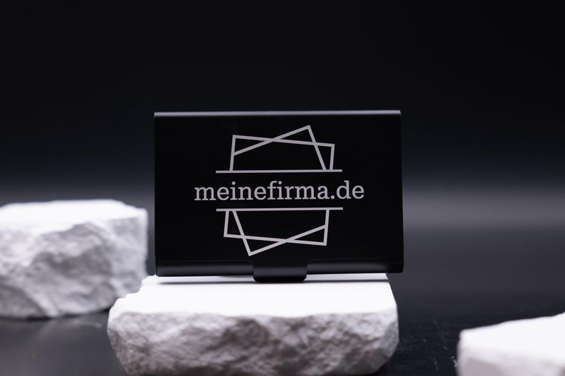 Schwarzes Visitenkartenetui aus Metall mit Lasergravur „meinefirma.de“ auf der Vorderseite. Modernes, minimalistisches Design auf weißem Stein fotografiert. Das Etui ist leicht, robust und perfekt für Geschäftsleute, die ihre Visitenkarten stilvoll und professionell präsentieren möchten. Die Gravur macht es zu einem personalisierten Accessoire für Büro, Meetings oder Networking.
