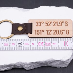 Personalisierter Holz Schlüsselanhänger mit gravierten Koordinaten „33° 52' 21.9'' S, 151° 12' 20.6'' O“ wird mit Maßband präsentiert. Das Maßband zeigt eine Länge von ca. 9 cm, wodurch der Anhänger handlich und stabil ist. Helles Holz kombiniert mit braunem Lederband und Metallring in Antik-Gold sorgt für edlen Look. Perfekt für individuelle Gravuren, Koordinaten oder Namen, ein ideales Geschenk für Partner und Reisende.