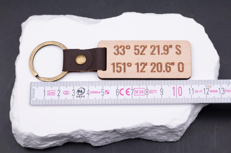 Personalisierter Holz Schlüsselanhänger mit gravierten Koordinaten „33° 52' 21.9'' S, 151° 12' 20.6'' O“ wird mit Maßband präsentiert. Das Maßband zeigt eine Länge von ca. 9 cm, wodurch der Anhänger handlich und stabil ist. Helles Holz kombiniert mit braunem Lederband und Metallring in Antik-Gold sorgt für edlen Look. Perfekt für individuelle Gravuren, Koordinaten oder Namen, ein ideales Geschenk für Partner und Reisende.