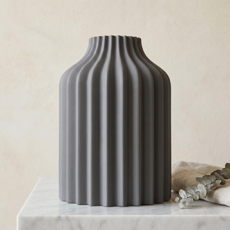 Może przedstawiać: Szary ceramiczny wazon z żebrowanym wzorem. Wazon ma cylindryczny korpus i lekko rozszerzoną g&oacute;rę. Stoi na białej marmurowej powierzchni. Gałązka eukaliptusa spoczywa na lnianej szmatce obok wazonu.