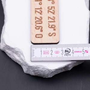 Nahaufnahme eines gravierten Holz Schlüsselanhängers mit Maßband. Das Maßband zeigt eine Breite von ca. 3 cm, wodurch der Anhänger kompakt und robust wirkt. Gravur mit Koordinaten macht ihn zu einem einzigartigen Accessoire. Gefertigt aus hellem Holz mit braunem Lederband und stabilem Metallring in Antik-Gold. Ideal für personalisierte Gravuren von Namen, Orten oder Sprüchen, ein besonderes Geschenk mit praktischem Nutzen.