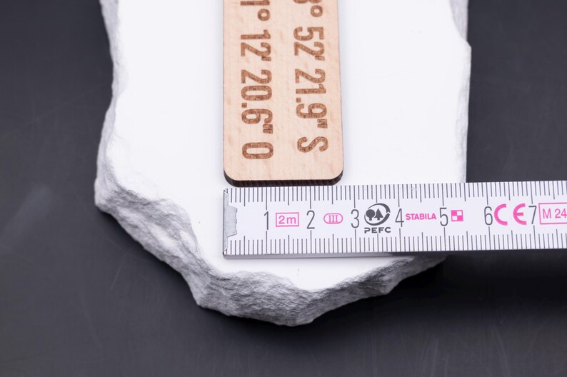 Nahaufnahme eines gravierten Holz Schlüsselanhängers mit Maßband. Das Maßband zeigt eine Breite von ca. 3 cm, wodurch der Anhänger kompakt und robust wirkt. Gravur mit Koordinaten macht ihn zu einem einzigartigen Accessoire. Gefertigt aus hellem Holz mit braunem Lederband und stabilem Metallring in Antik-Gold. Ideal für personalisierte Gravuren von Namen, Orten oder Sprüchen, ein besonderes Geschenk mit praktischem Nutzen.