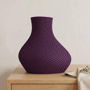 May include: A deep purple vase with a textured, lattice-like pattern. The vase has a wide base that tapers to a narrow neck and flared opening. The vase is sitting on a light-colored wooden surface.