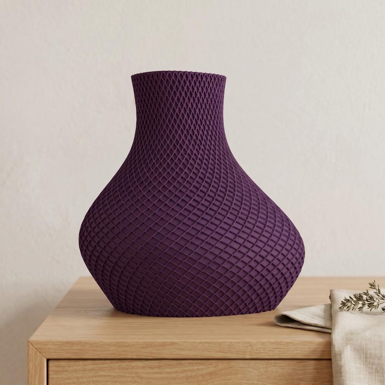 May include: A deep purple vase with a textured, lattice-like pattern. The vase has a wide base that tapers to a narrow neck and flared opening. The vase is sitting on a light-colored wooden surface.