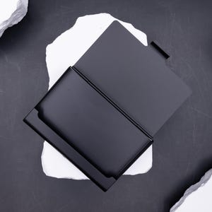 Geöffnetes Visitenkartenetui aus schwarzem Metall, von oben auf weißem Stein fotografiert. Innenraum bietet Platz für mehrere Karten, robust und langlebig. Das Etui schützt Visitenkarten oder Kreditkarten zuverlässig und passt dank seines leichten Gewichts in jede Tasche. Funktionalität kombiniert mit elegantem, minimalistischem Design – ideal für den professionellen Auftritt.