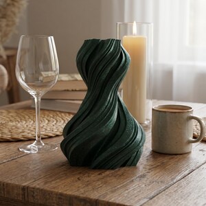 K&ouml;nnte beinhalten: Eine dunkelgr&uuml;ne Vase mit strukturiertem, wirbelndem Design. Ein Weinglas, eine brennende Kerze in einem Glasbeh&auml;lter und eine Kaffeetasse stehen auf einem Holztisch. Die Vase hat eine einzigartige, k&uuml;nstlerische Form.