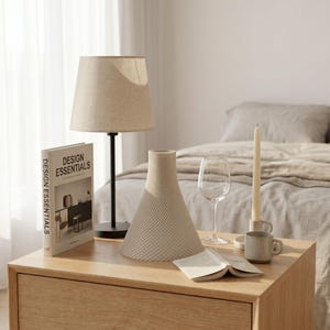 Peut inclure: Un abat-jour beige sur une base de lampe noire est pos&eacute; sur une table de chevet en bois. Un vase beige textur&eacute;, un verre &agrave; vin, une bougie, une tasse et un livre intitul&eacute; "DESIGN ESSENTIALS" sont &eacute;galement sur la table. Un lit est en arri&egrave;re-plan.
