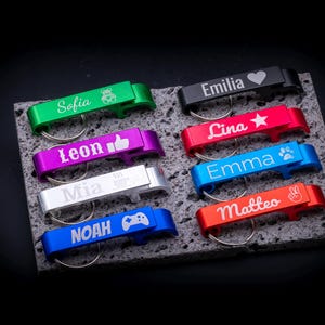 Personalisierbare Flaschenöffner Schlüsselanhänger aus Aluminium in verschiedenen Farben mit Gravur von Namen und Symbolen. Abgebildet sind Modelle in Grün, Schwarz, Rot, Blau, Silber, Lila und Orange mit Gravuren wie Herz, Stern, Bärchen, Pfote, Daumen hoch oder Spielecontroller. Praktisch als Schlüsselanhänger mit integriertem Kapselheber und individueller Gravur – ideal als Geschenk oder Werbeartikel.