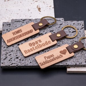 Drei personalisierte Schlüsselanhänger aus Holz mit Kunstlederband und Messingring, graviert mit individuellen Sprüchen: „Toms Abenteuerbund“, „Opa’s Bastelbude“ und „Papas Schlüsselreich“ mit Herz-Symbol. Die rechteckigen Anhänger liegen dekorativ auf Natursteinplatten und verbinden rustikalen Holzcharakter mit modernem Design. Ideal als Geschenk für Vater, Opa oder Kinder, praktisch und stilvoll zugleich.