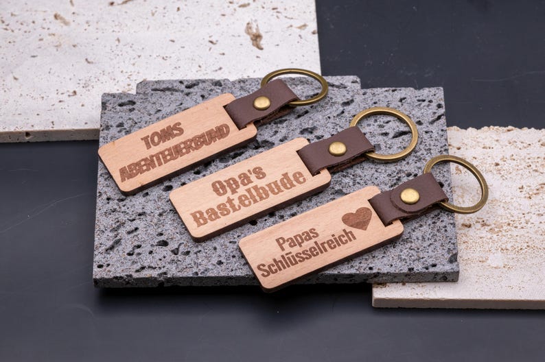 Drei personalisierte Schlüsselanhänger aus Holz mit Kunstlederband und Messingring, graviert mit individuellen Sprüchen: „Toms Abenteuerbund“, „Opa’s Bastelbude“ und „Papas Schlüsselreich“ mit Herz-Symbol. Die rechteckigen Anhänger liegen dekorativ auf Natursteinplatten und verbinden rustikalen Holzcharakter mit modernem Design. Ideal als Geschenk für Vater, Opa oder Kinder, praktisch und stilvoll zugleich.