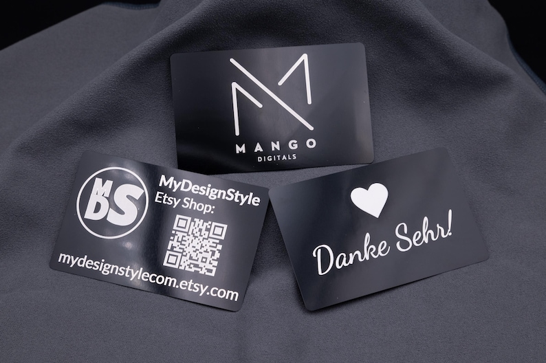 Drei schwarze Visitenkarten aus Aluminium mit matter Oberfläche und weißer Gravur, auf grauem Stoff präsentiert. Links das MDS-Shoplogo mit QR-Code, in der Mitte das Mango-Digitals-Logo und rechts eine Dankeskarte mit Herzsymbol und Schriftzug „Danke Sehr!“. Die Karten sind robust, im Kreditkartenformat (8,6 × 5,4 cm), langlebig und wirken modern und exklusiv. Perfekt für Business, Branding, Networking und kreative Kundendankesgesten.