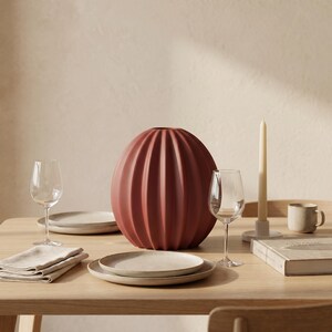 May include: A wooden dining table set with a large, terracotta-coloured, ribbed vase as a centrepiece. The table also features wine glasses, plates, a candle, a mug, and a folded napkin. The setting has a minimalist design and a warm, neutral colour scheme.