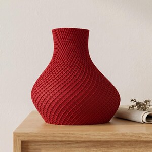 May include: A red decorative vase with a textured, lattice-like pattern. The vase has a wide base that tapers to a narrow neck and a flared top. The vase is sitting on a wooden surface.