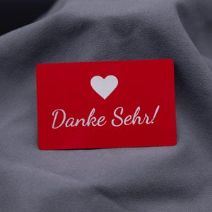 Einzelne rote Dankeskarte aus Aluminium mit eingraviertem Herzsymbol und Schriftzug „Danke Sehr!“, auf grauem Stoff präsentiert. Die Metallkarte hat eine matte Oberfläche, ist langlebig, stabil und wirkt besonders hochwertig. Perfekt als exklusive Dankeskarte für Kunden oder Partner und eine stilvolle Alternative zu Papierkarten.