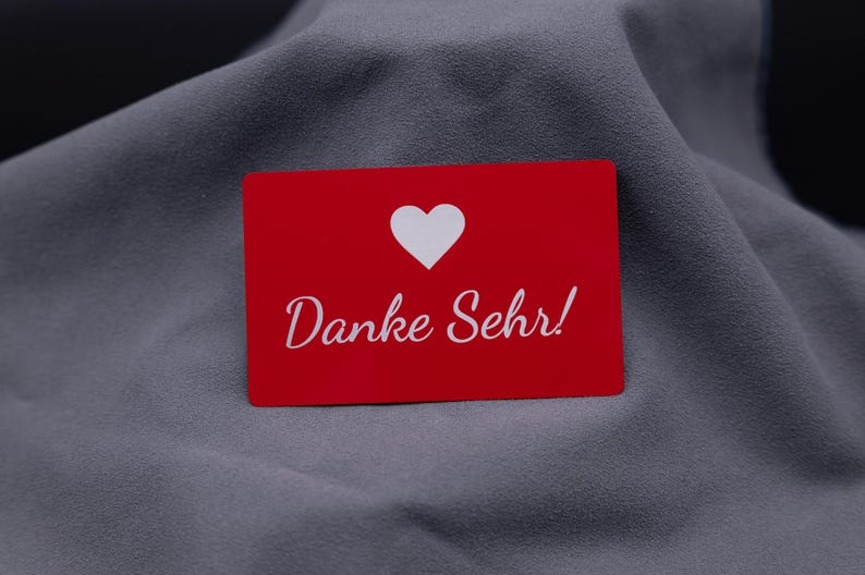Einzelne rote Dankeskarte aus Aluminium mit eingraviertem Herzsymbol und Schriftzug „Danke Sehr!“, auf grauem Stoff präsentiert. Die Metallkarte hat eine matte Oberfläche, ist langlebig, stabil und wirkt besonders hochwertig. Perfekt als exklusive Dankeskarte für Kunden oder Partner und eine stilvolle Alternative zu Papierkarten.