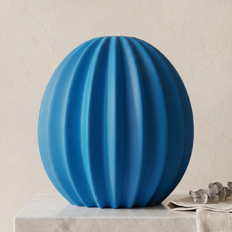 May include: A striking blue spherical vase with vertical ribbed detailing. The vase sits on a white marble surface, highlighting its unique texture and shape. A stylish home decor accent.