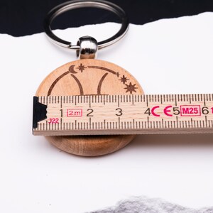 Detailaufnahme eines Holz-Schlüsselanhängers mit Gravur, zusammen mit einem Maßband zur Größenangabe. Der Anhänger zeigt das astrologische Symbol des Schützen, eingerahmt von Sternen und Mondsichel. Das Maßband verdeutlicht den Durchmesser von rund 4 cm. Der Schlüsselring ist aus Metall gefertigt. Diese Darstellung hebt die exakten Maße und die feine Gravur hervor, ideal zur Veranschaulichung der Produktdetails für Käufer.