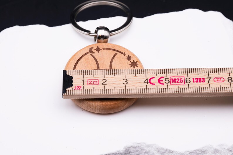 Detailaufnahme eines Holz-Schlüsselanhängers mit Gravur, zusammen mit einem Maßband zur Größenangabe. Der Anhänger zeigt das astrologische Symbol des Schützen, eingerahmt von Sternen und Mondsichel. Das Maßband verdeutlicht den Durchmesser von rund 4 cm. Der Schlüsselring ist aus Metall gefertigt. Diese Darstellung hebt die exakten Maße und die feine Gravur hervor, ideal zur Veranschaulichung der Produktdetails für Käufer.
