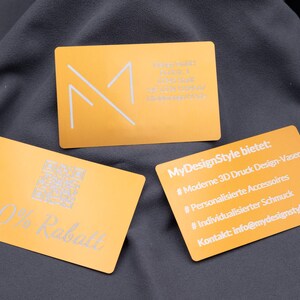 Rückseiten von drei Aluminium-Visitenkarten in Hellgold. Eine Karte zeigt „20% Rabatt“ mit QR-Code, eine weitere listet MyDesignStyle-Angebote wie 3D-Druck Vasen, Schmuck und Accessoires mit Kontakt-E-Mail, daneben eine Firmenkarte von Mango GmbH mit Adresse, Telefonnummer und Mail. Die gravierten Metallkarten sind stabil, langlebig und ideal für professionelle Werbung oder personalisierte Marketingaktionen.