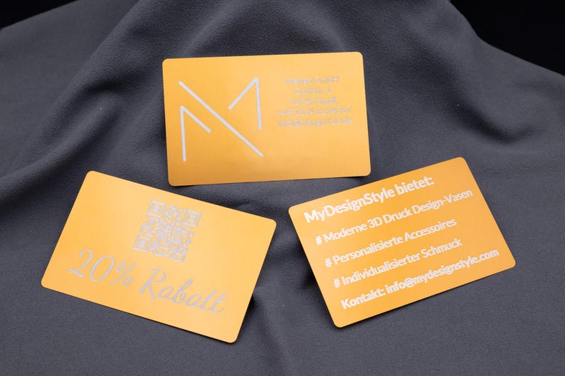 Rückseiten von drei Aluminium-Visitenkarten in Hellgold. Eine Karte zeigt „20% Rabatt“ mit QR-Code, eine weitere listet MyDesignStyle-Angebote wie 3D-Druck Vasen, Schmuck und Accessoires mit Kontakt-E-Mail, daneben eine Firmenkarte von Mango GmbH mit Adresse, Telefonnummer und Mail. Die gravierten Metallkarten sind stabil, langlebig und ideal für professionelle Werbung oder personalisierte Marketingaktionen.