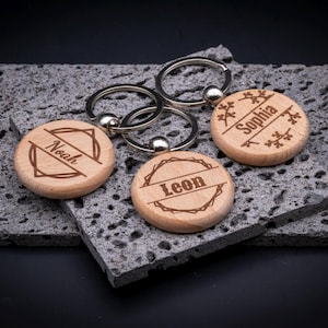 Runder Schlüsselanhänger aus Holz mit individueller Gravur. Drei Beispiele: „Noah“ mit geometrischem Rahmen, „Leon“ mit rustikalem Kranzmotiv und „Sophia“ mit verspielten Ästen. Edler Holzanhänger mit Metallring, ideal als personalisiertes Geschenk.