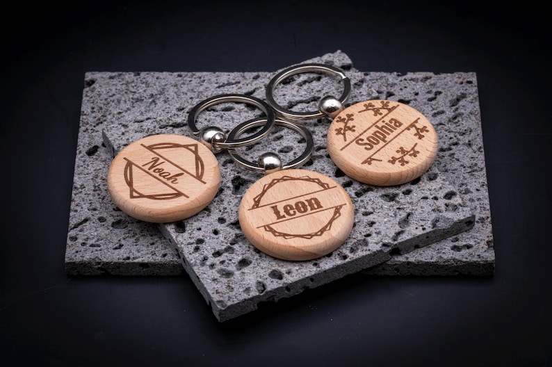 Runder Schlüsselanhänger aus Holz mit individueller Gravur. Drei Beispiele: „Noah“ mit geometrischem Rahmen, „Leon“ mit rustikalem Kranzmotiv und „Sophia“ mit verspielten Ästen. Edler Holzanhänger mit Metallring, ideal als personalisiertes Geschenk.