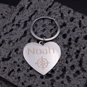 Schlüsselanhänger Herz aus Edelstahl mit Gravur „Noah“ und Kompasssymbol. Die Gravur steht für Orientierung, Freundschaft und Liebe. Der glänzende Metallanhänger ruht auf dunklen, strukturierten Steinen, was den Kontrast und die edle Gravur betont. Perfektes Geschenk für Reisefans, Partner oder beste Freunde.