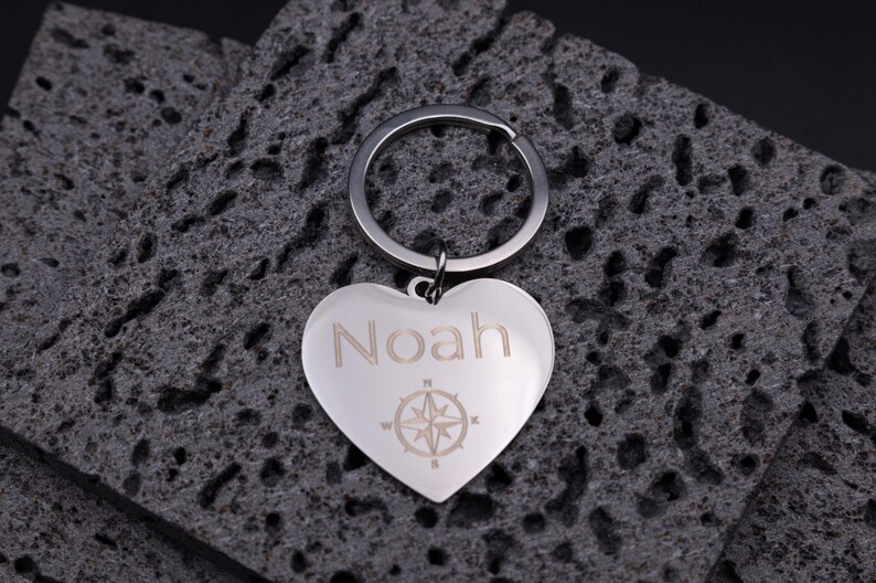 Schlüsselanhänger Herz aus Edelstahl mit Gravur „Noah“ und Kompasssymbol. Die Gravur steht für Orientierung, Freundschaft und Liebe. Der glänzende Metallanhänger ruht auf dunklen, strukturierten Steinen, was den Kontrast und die edle Gravur betont. Perfektes Geschenk für Reisefans, Partner oder beste Freunde.